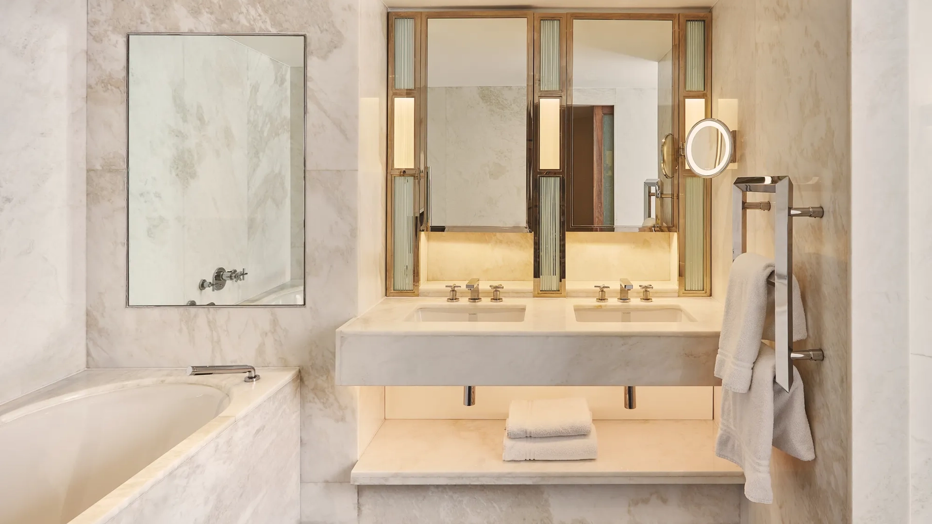 Marble bathroom with a bathtub, double vanity, illuminated mirrors, towel rack, and folded white towels on a shelf beneath the sink.