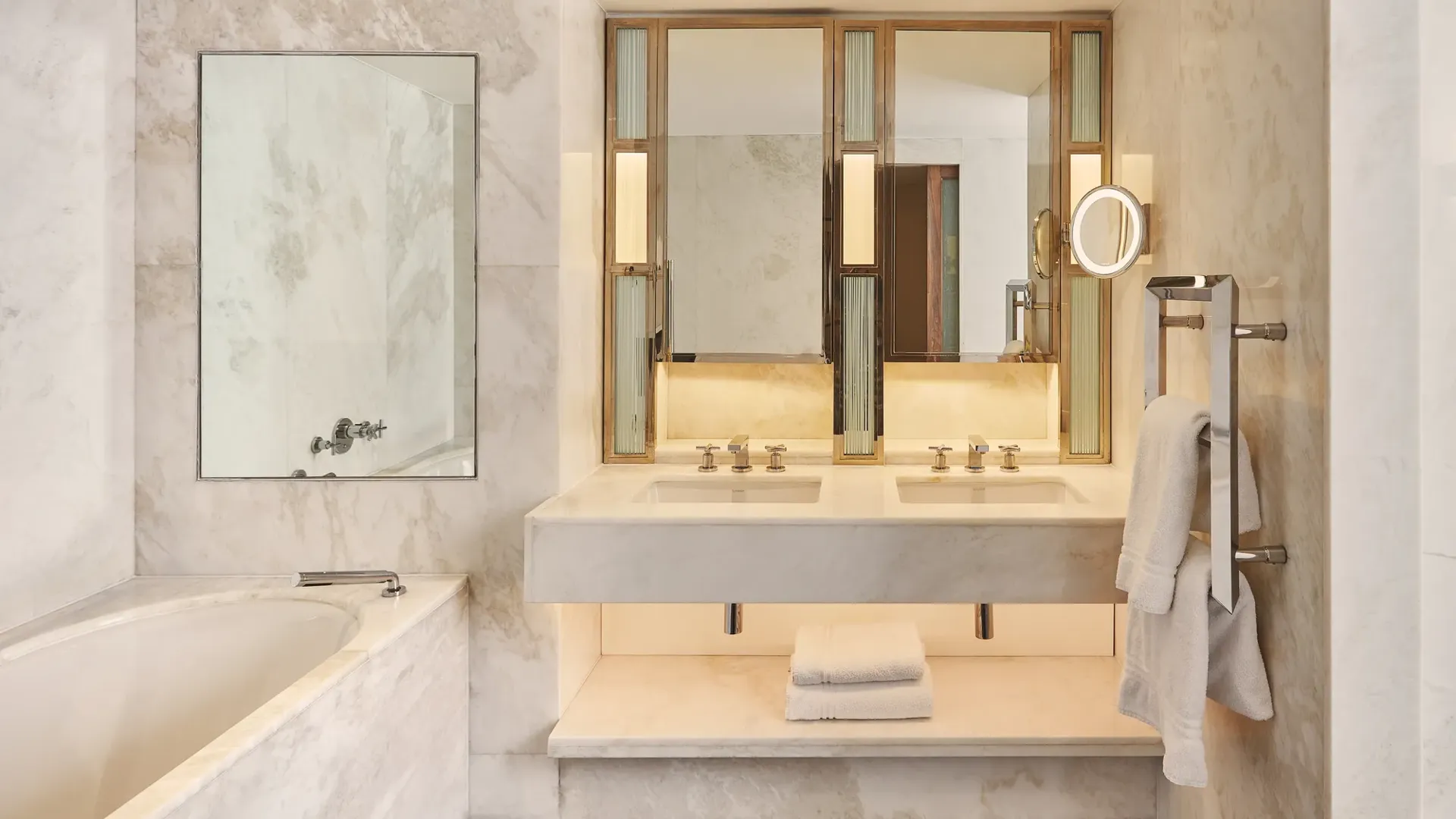 Marble bathroom with a bathtub, double vanity, illuminated mirrors, towel rack, and folded white towels on a shelf beneath the sink.