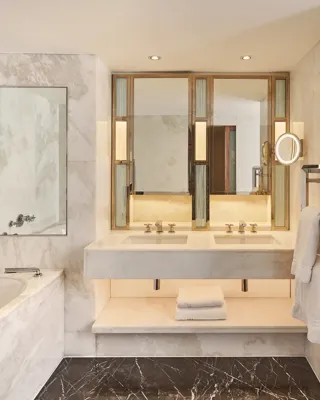Marble bathroom with a bathtub, double vanity, illuminated mirrors, towel rack, and folded white towels on a shelf beneath the sink.