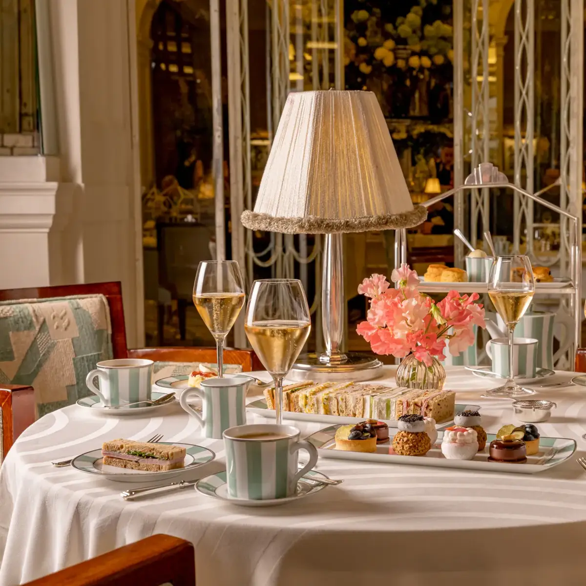 Afternoon tea table with champagne, sandwiches and pastries around a lamp centrepiece in an elegant restaurant.