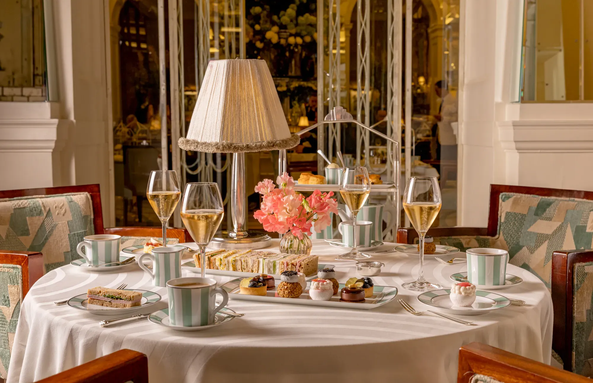 Afternoon tea table with champagne, sandwiches and pastries around a lamp centrepiece in an elegant restaurant.