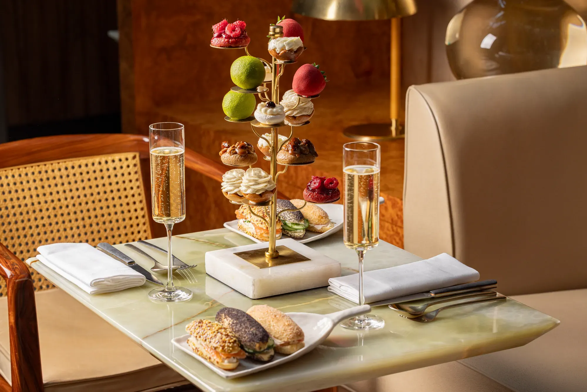 Tiered stand of pastries with champagne flutes and savoury eclairs on marble table.