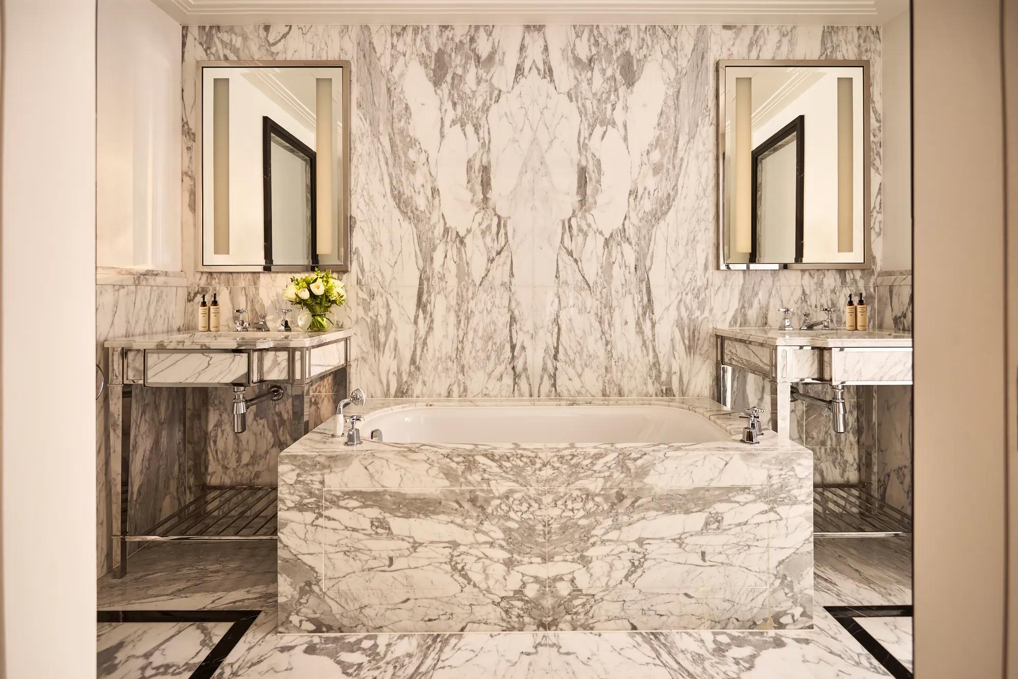 Marble bathroom with a freestanding bathtub, twin vanity sinks, mirrored walls, and grey veined stone extending across floors and surfaces.