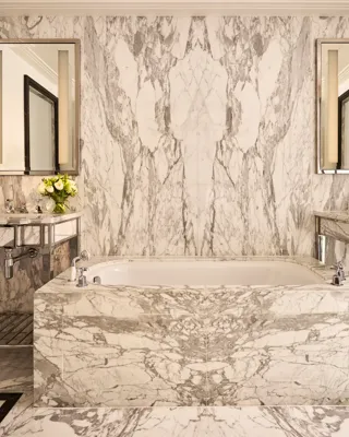 Marble bathroom with a freestanding bathtub, twin vanity sinks, mirrored walls, and grey veined stone extending across floors and surfaces.