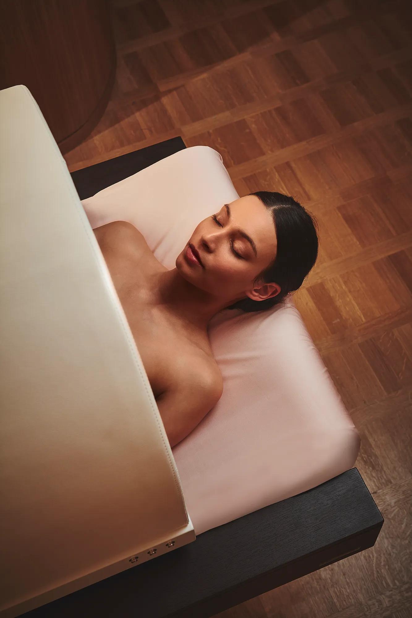 Guest lying on a treatment bed during a spa experience, eyes closed, partially covered by a blanket in a calm, wood-floored room.