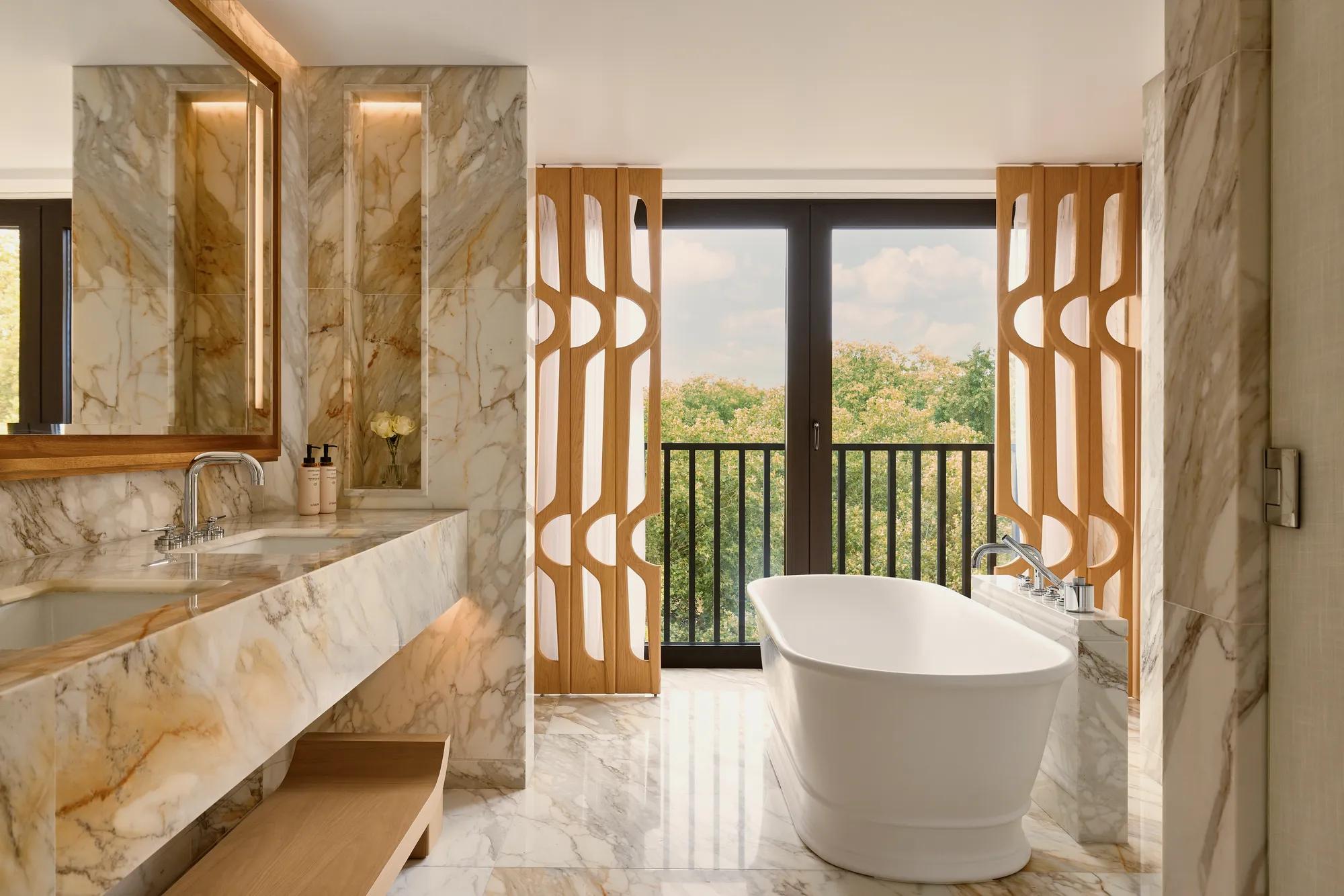 Marble bathroom with freestanding bathtub, dual sinks, and large windows with leafy park views