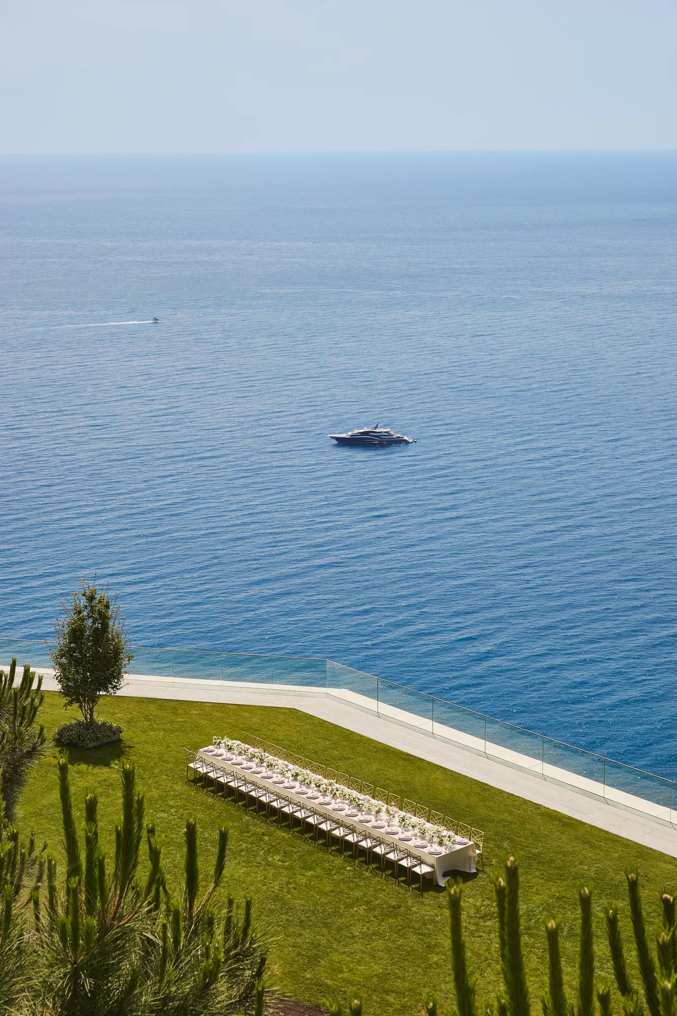 Outdoor event setup with long banquet table on a lawn overlooking the sea, with a yacht visible on the water.