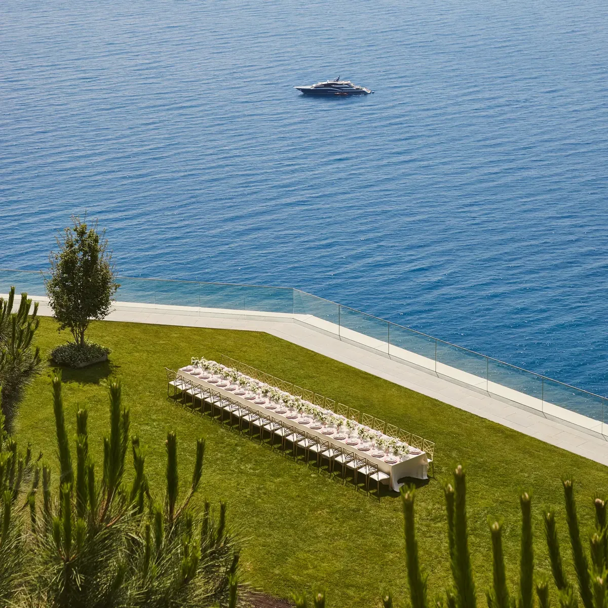 Installation extérieure avec longue table de banquet sur pelouse surplombant la mer, avec un yacht visible au large.