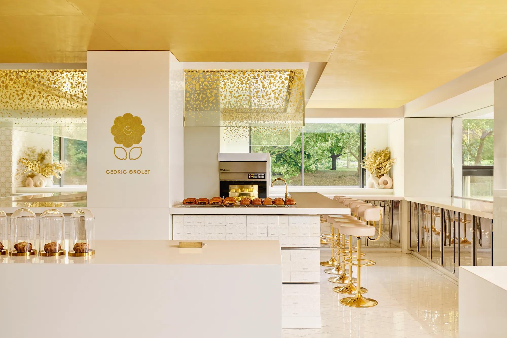 Modern patisserie counter with gold accents, displaying pastries under glass domes and rows of brioche buns, with seating along the side.
