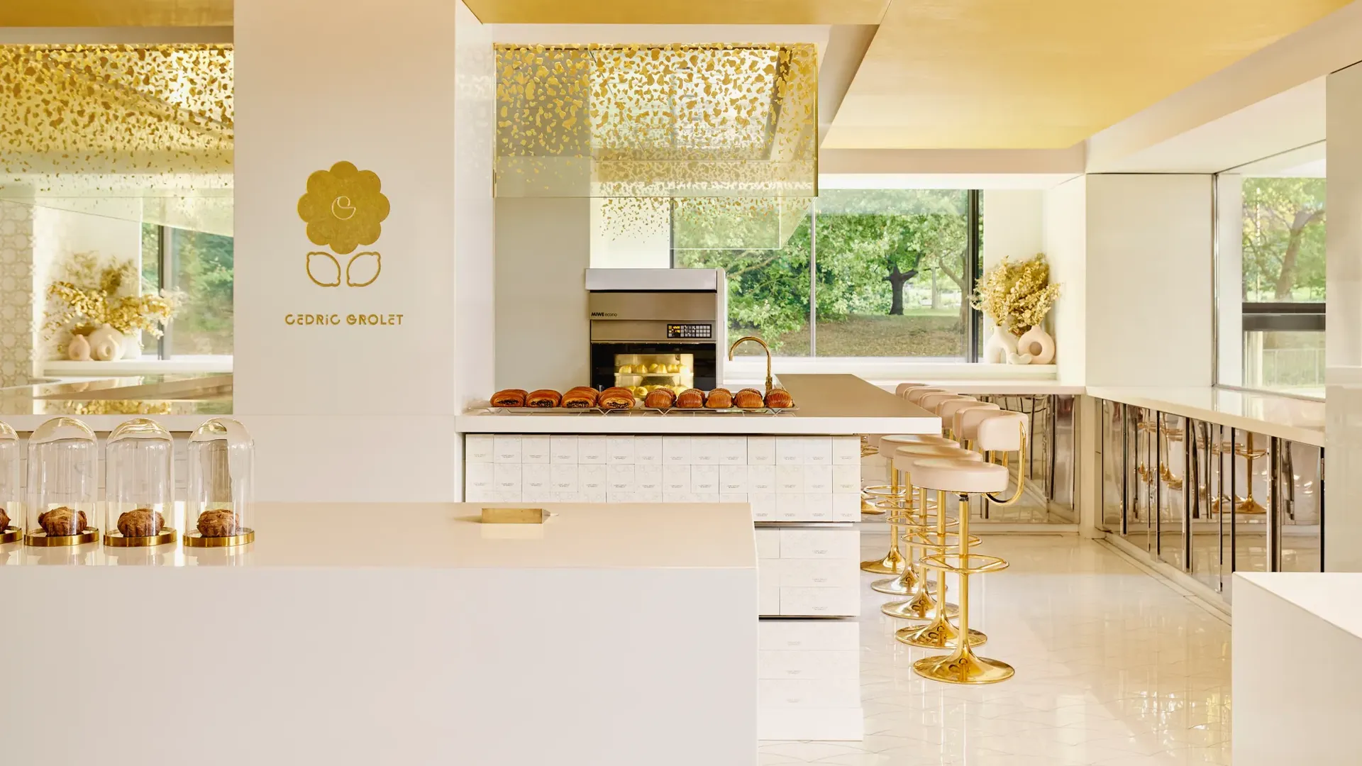 Modern patisserie counter with gold accents, displaying pastries under glass domes and rows of brioche buns, with seating along the side.