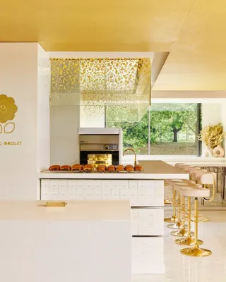Modern patisserie counter with gold accents, displaying pastries under glass domes and rows of brioche buns, with seating along the side.
