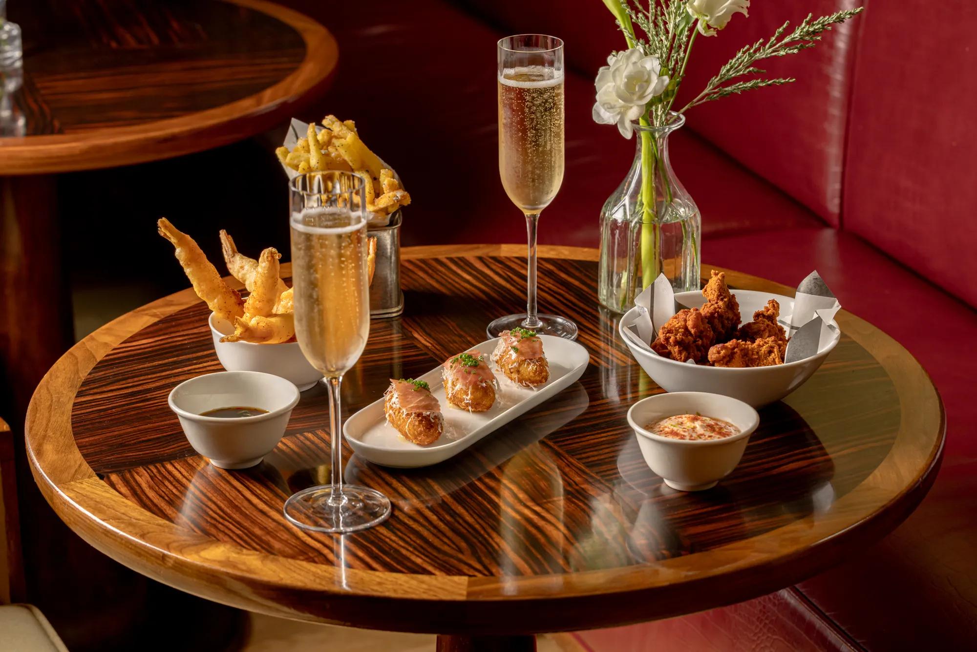 Champagne flutes with fried chicken, tempura, and salmon bites on a polished wood table with red leather seating