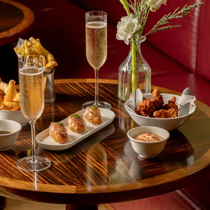 Champagne flutes with fried chicken, tempura, and salmon bites on a polished wood table with red leather seating