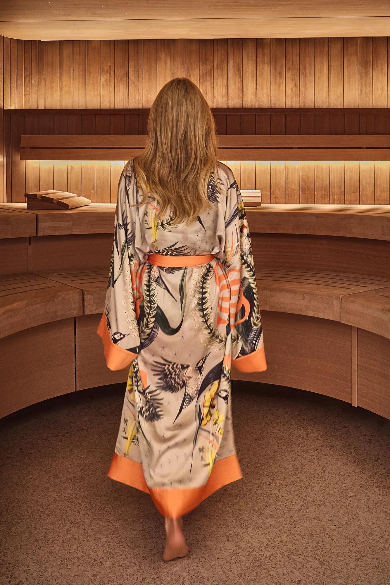 Person wearing patterned robe walking barefoot through a wooden spa interior with curved seating.