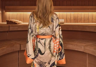 Person wearing patterned robe walking barefoot through a wooden spa interior with curved seating.