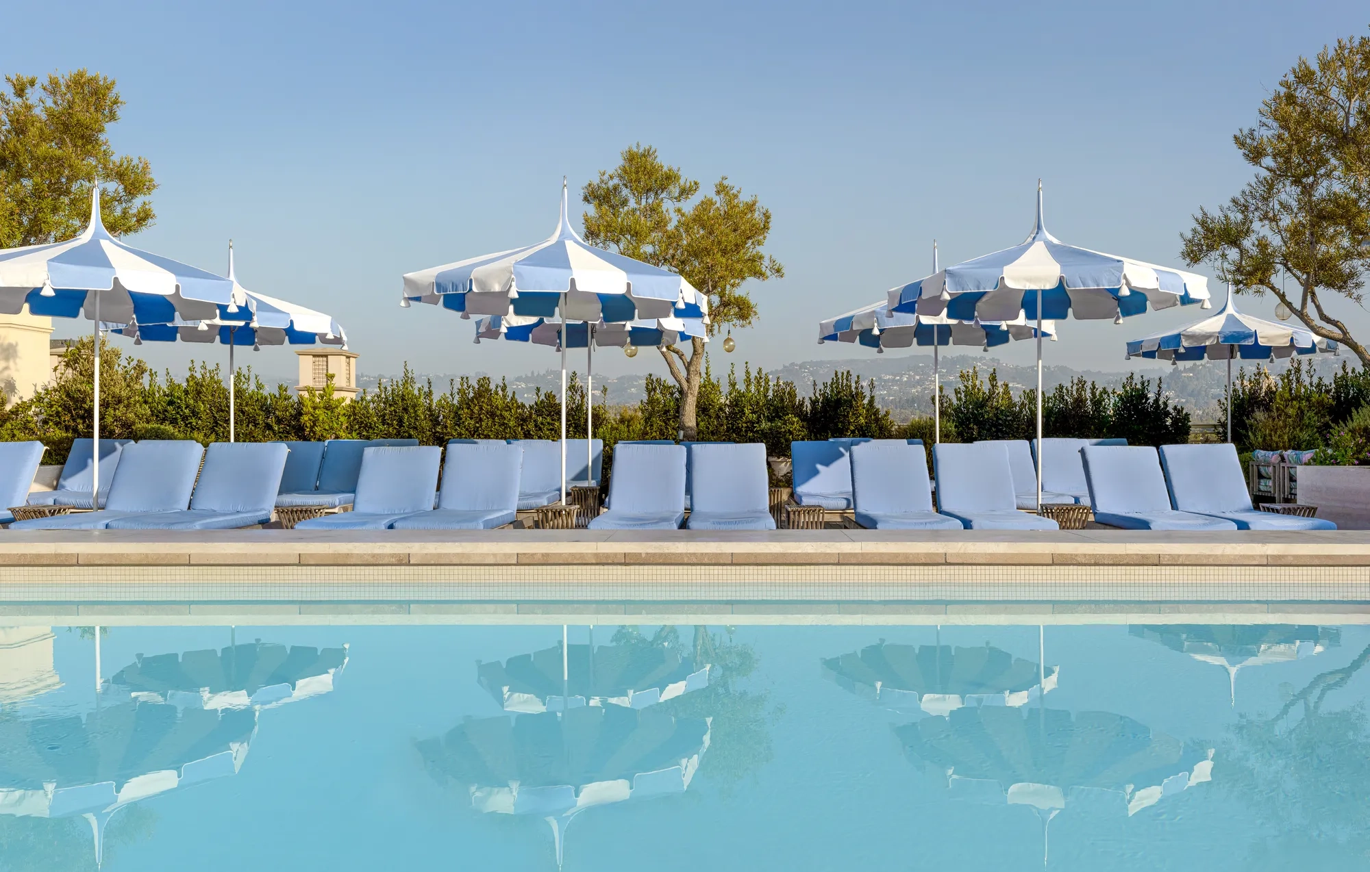 The Maybourne Beverly Hills ninth floor rooftop pool with blue loungers and striped umbrellas reflected in the water.