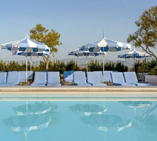 The Maybourne Beverly Hills ninth floor rooftop pool with blue loungers and striped umbrellas reflected in the water.