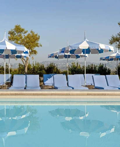 The Maybourne Beverly Hills ninth floor rooftop pool with blue loungers and striped umbrellas reflected in the water.