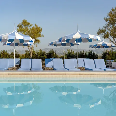 The Maybourne Beverly Hills ninth floor rooftop pool with blue loungers and striped umbrellas reflected in the water.