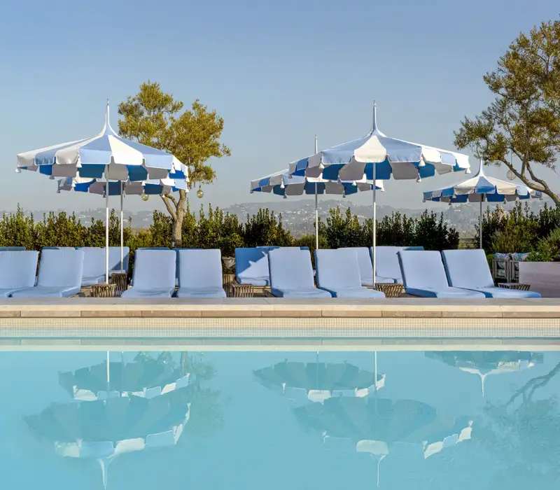 The Maybourne Beverly Hills ninth floor rooftop pool with blue loungers and striped umbrellas reflected in the water.