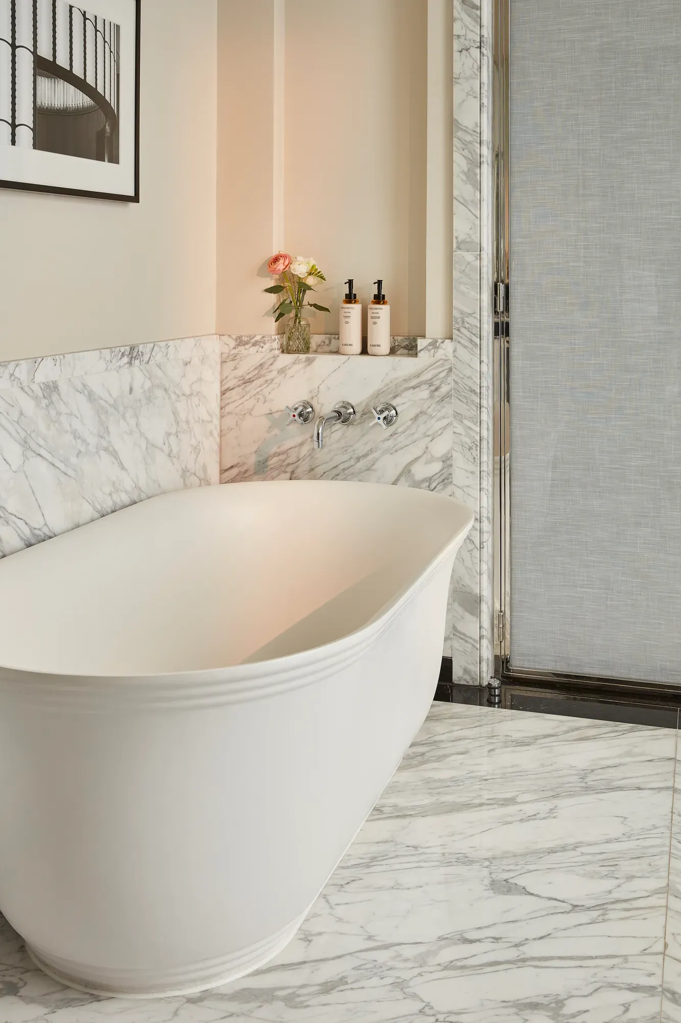 Marble bathroom with freestanding bathtub, wall-mounted taps, and toiletries on recessed shelf.
