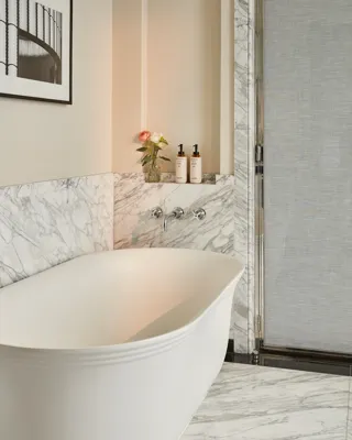 Marble bathroom with freestanding bathtub, wall-mounted taps, and toiletries on recessed shelf.