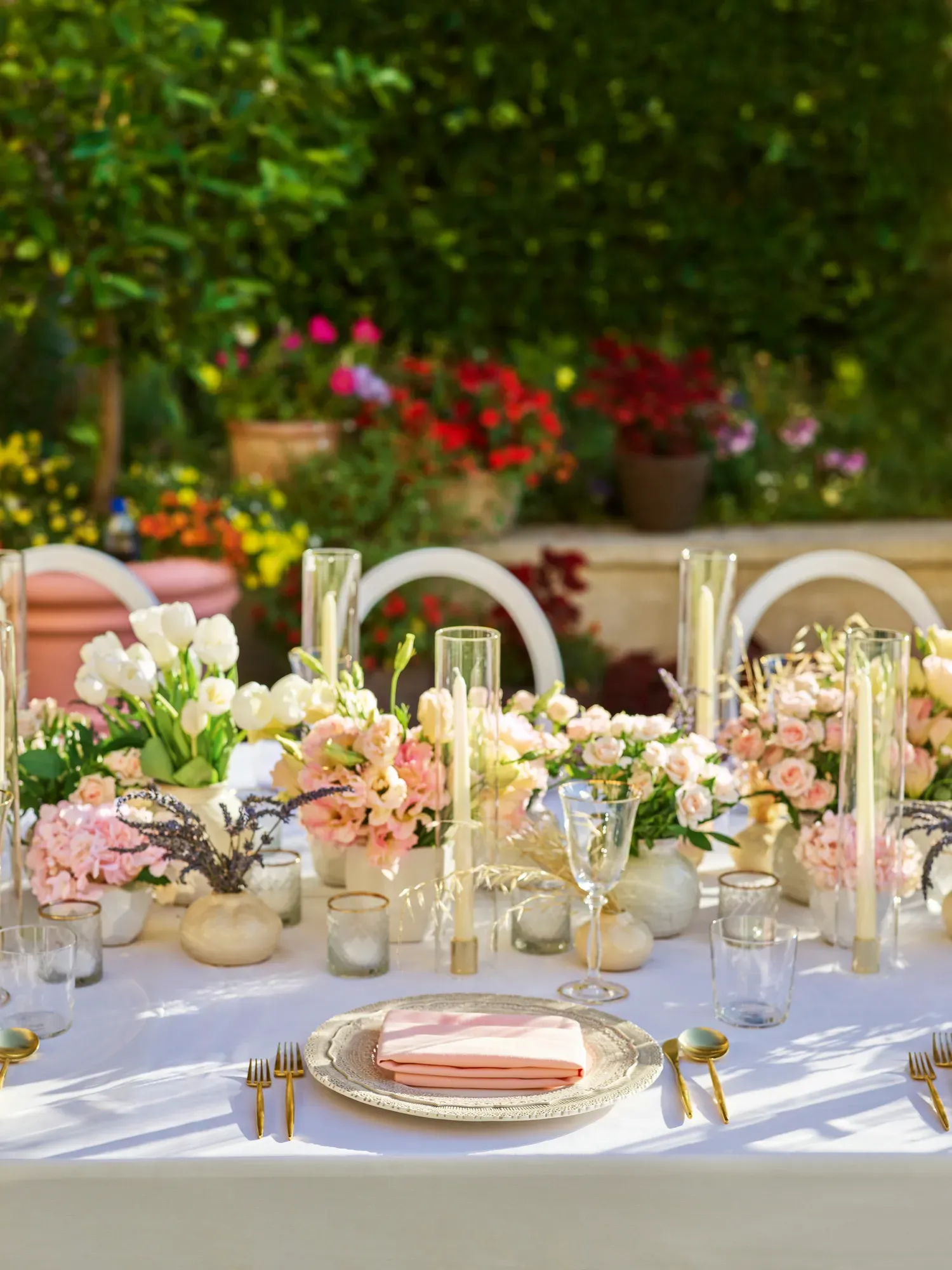 Place setting with pink napkin on textured charger amid pastel flowers, glass hurricanes, and gold-rim votives against a green hedge.