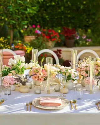 Place setting with pink napkin on textured charger amid pastel flowers, glass hurricanes, and gold-rim votives against a green hedge.