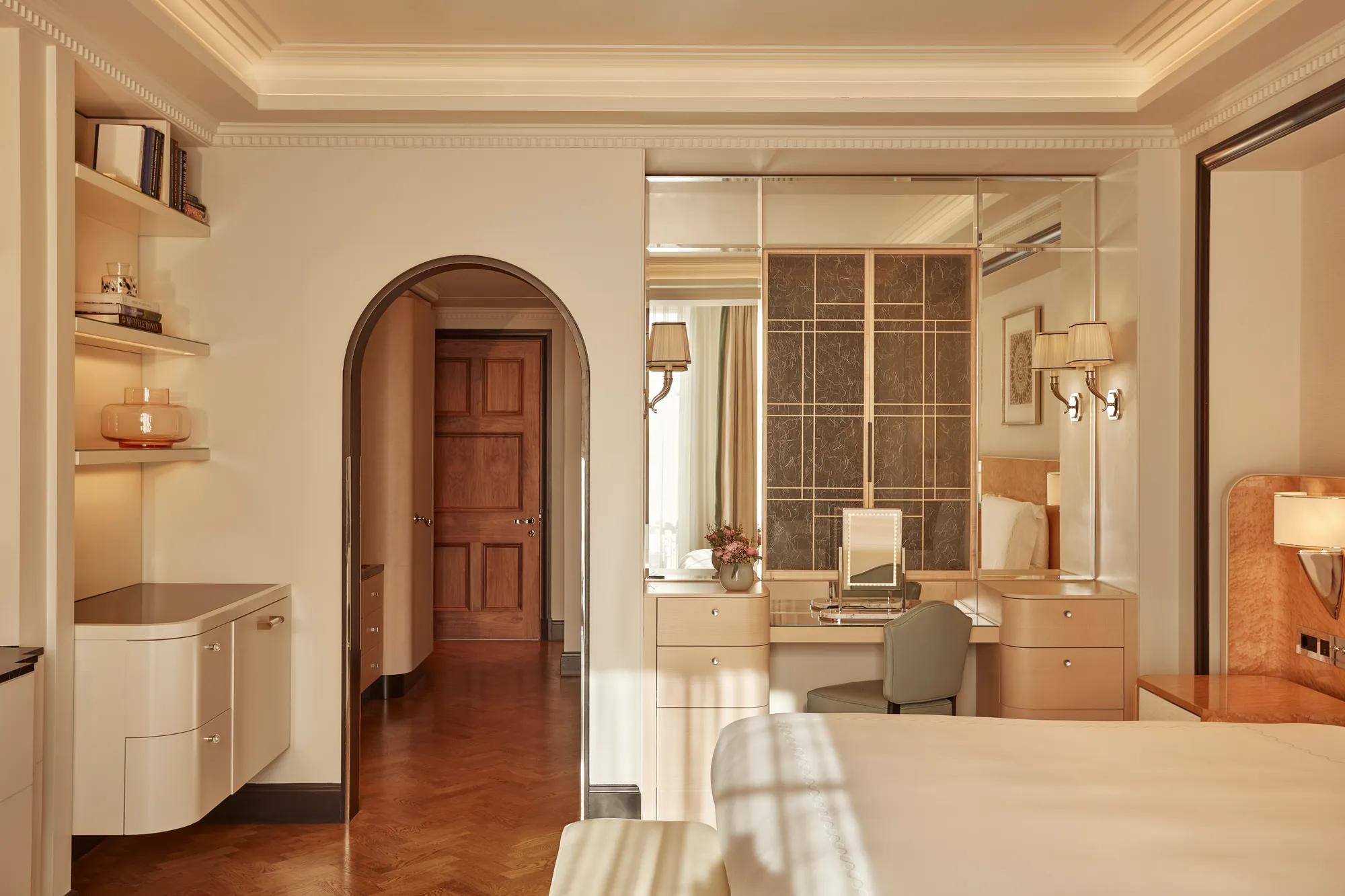 Elegant bedroom with light wood furnishings, mirrored vanity area, and arched doorway leading to a hallway, softly lit by wall sconces and natural light.