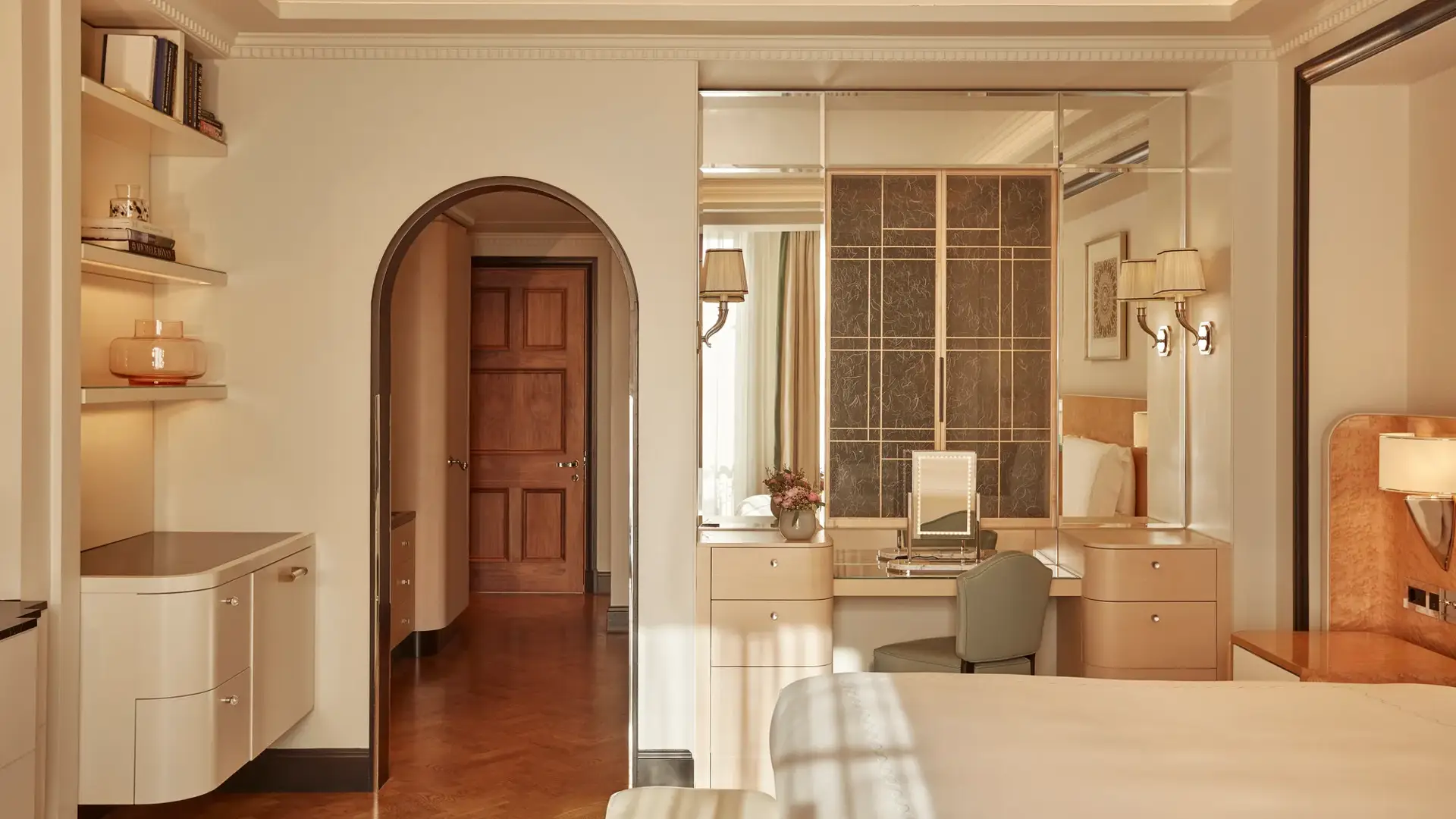 Elegant bedroom with light wood furnishings, mirrored vanity area, and arched doorway leading to a hallway, softly lit by wall sconces and natural light.