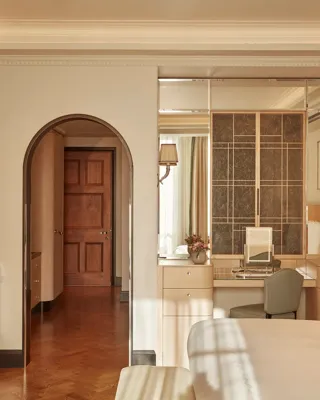 Elegant bedroom with light wood furnishings, mirrored vanity area, and arched doorway leading to a hallway, softly lit by wall sconces and natural light.