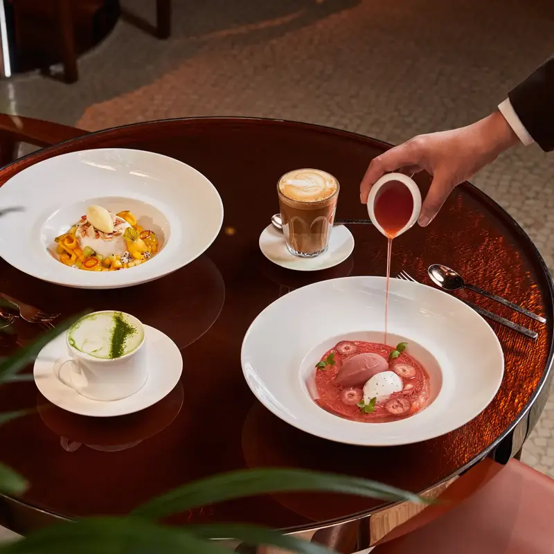 A person pouring red sauce into a dessert bowl beside another plated dish, latte, and matcha drink on a round table.