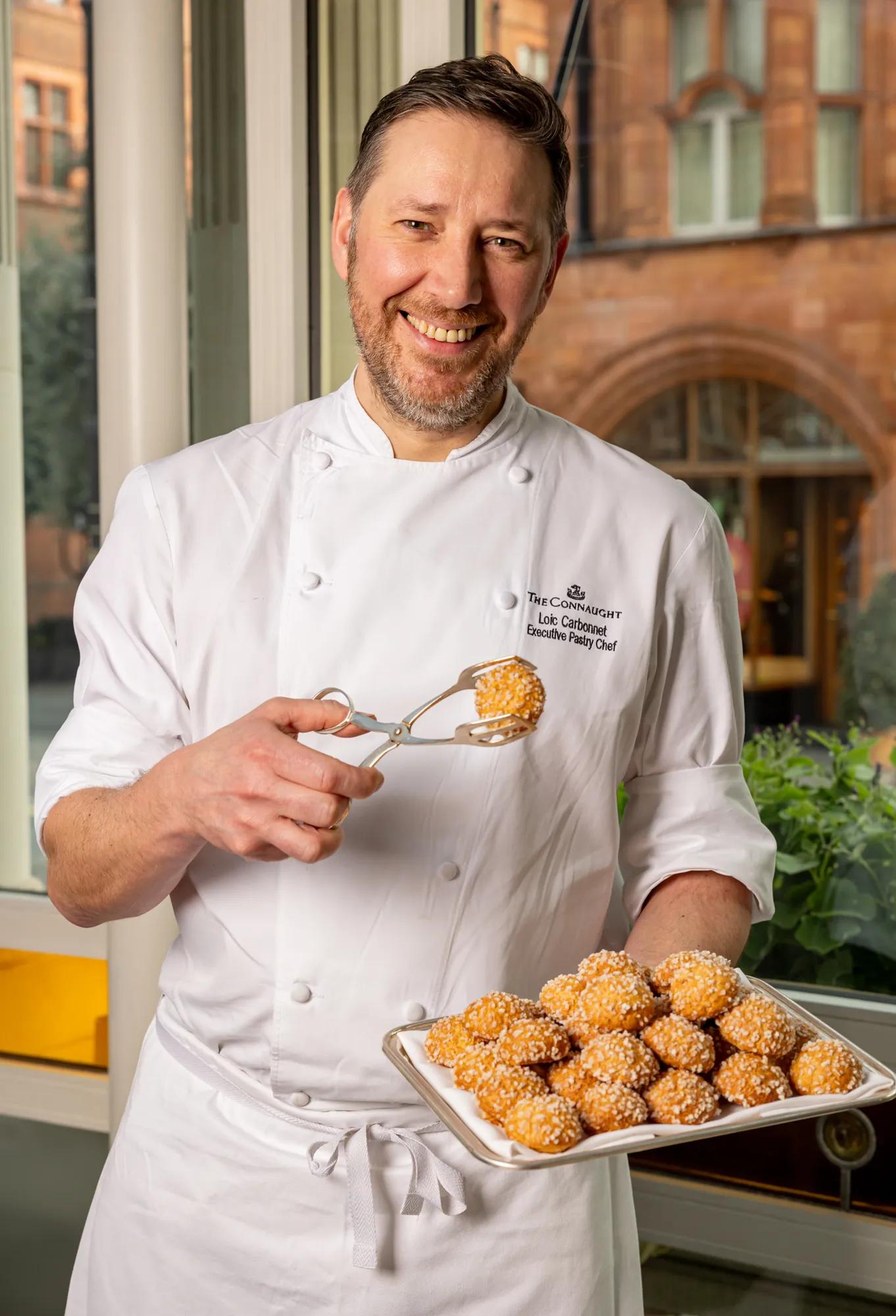 A picture of a chef in white uniform holding a plate of pastries and a pastry in one hand