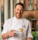 A picture of a chef in white uniform holding a plate of pastries and a pastry in one hand