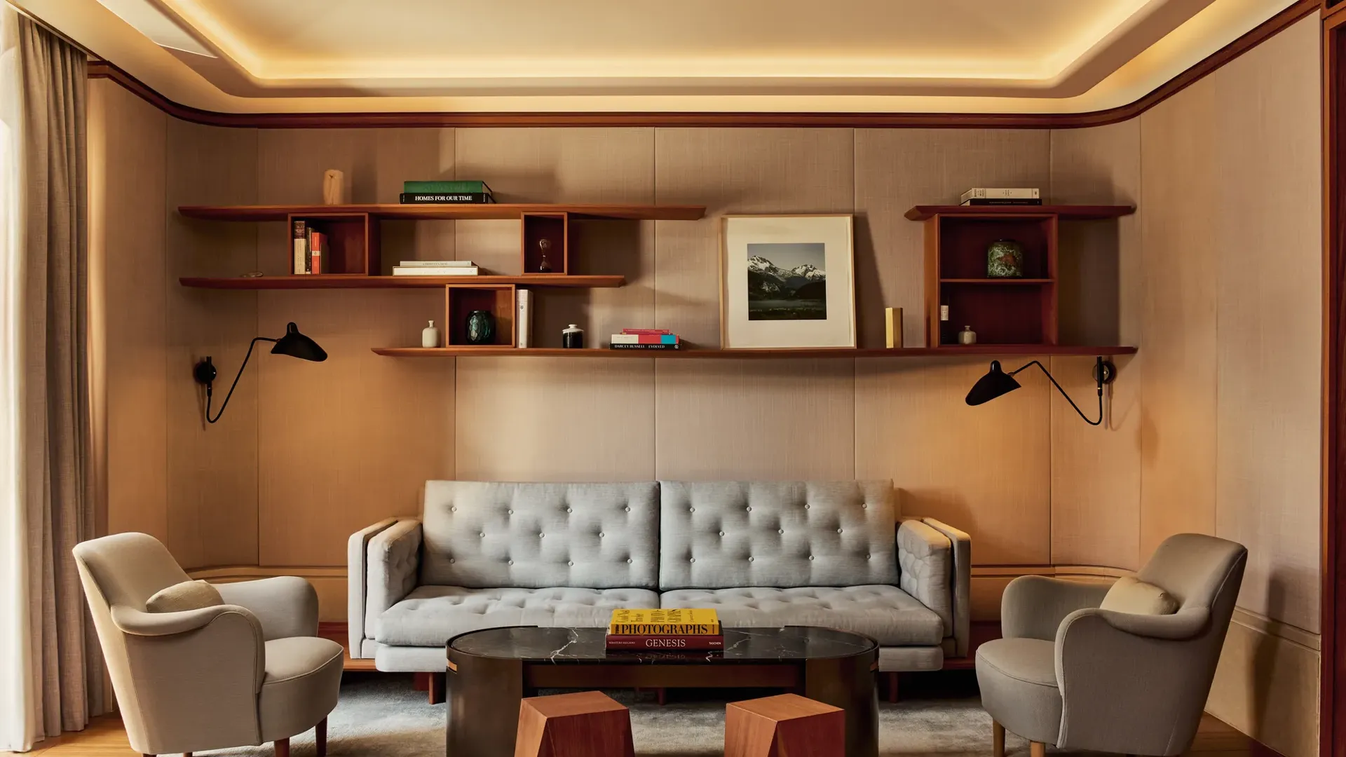 Elegant living room in the Knightsbridge Suite featuring a tufted grey sofa, two cream armchairs, and a black marble coffee table with geometric wooden stools, framed by wood shelving with books and art.