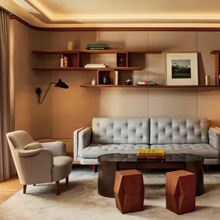 Elegant living room in the Knightsbridge Suite featuring a tufted grey sofa, two cream armchairs, and a black marble coffee table with geometric wooden stools, framed by wood shelving with books and art.