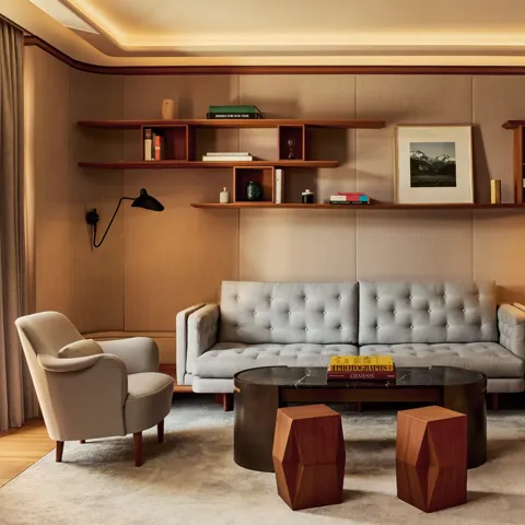 Elegant living room in the Knightsbridge Suite featuring a tufted grey sofa, two cream armchairs, and a black marble coffee table with geometric wooden stools, framed by wood shelving with books and art.
