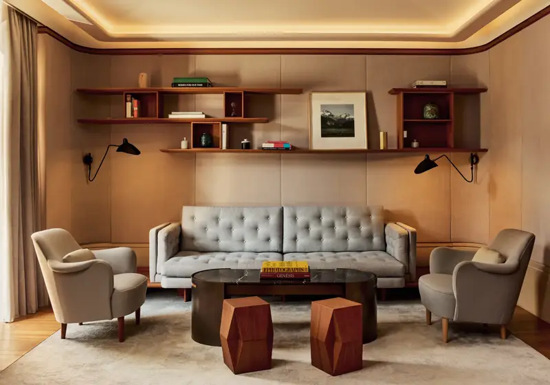 Elegant living room in the Knightsbridge Suite featuring a tufted grey sofa, two cream armchairs, and a black marble coffee table with geometric wooden stools, framed by wood shelving with books and art.