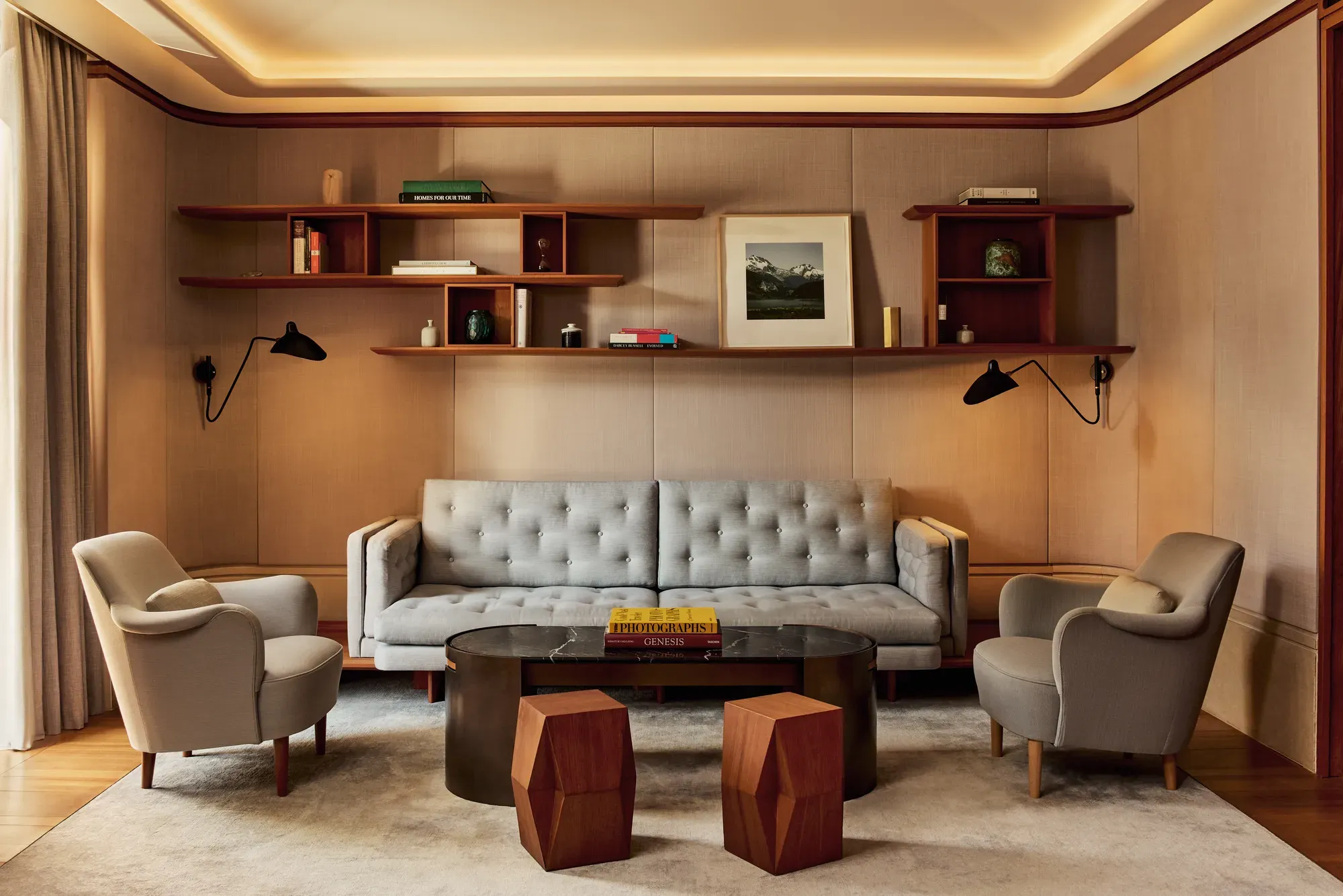Elegant living room in the Knightsbridge Suite featuring a tufted grey sofa, two cream armchairs, and a black marble coffee table with geometric wooden stools, framed by wood shelving with books and art.