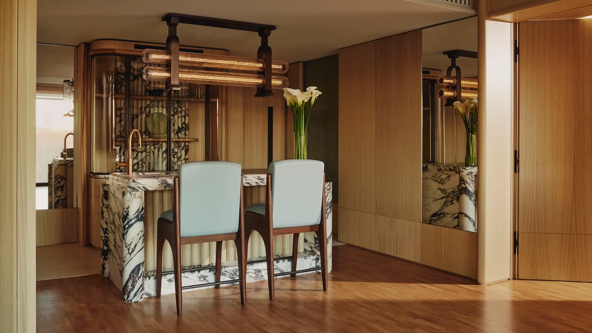 Elegant suite dining area with a marble-topped bar, two upholstered chairs, wood-paneled walls, mirrored accents, and modern pendant lighting, decorated with a tall vase of white flowers.