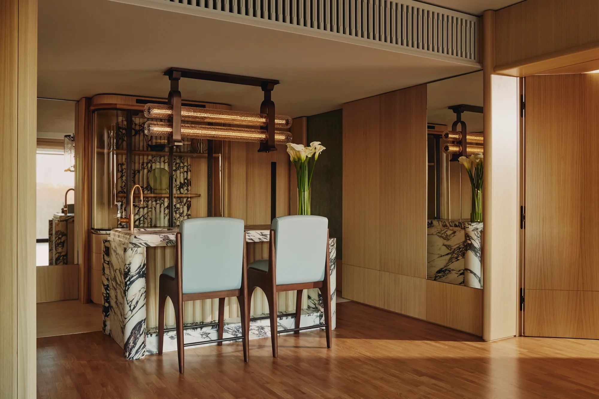 Elegant suite dining area with a marble-topped bar, two upholstered chairs, wood-paneled walls, mirrored accents, and modern pendant lighting, decorated with a tall vase of white flowers.