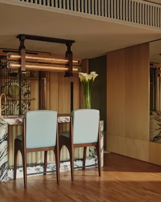 Elegant suite dining area with a marble-topped bar, two upholstered chairs, wood-paneled walls, mirrored accents, and modern pendant lighting, decorated with a tall vase of white flowers.