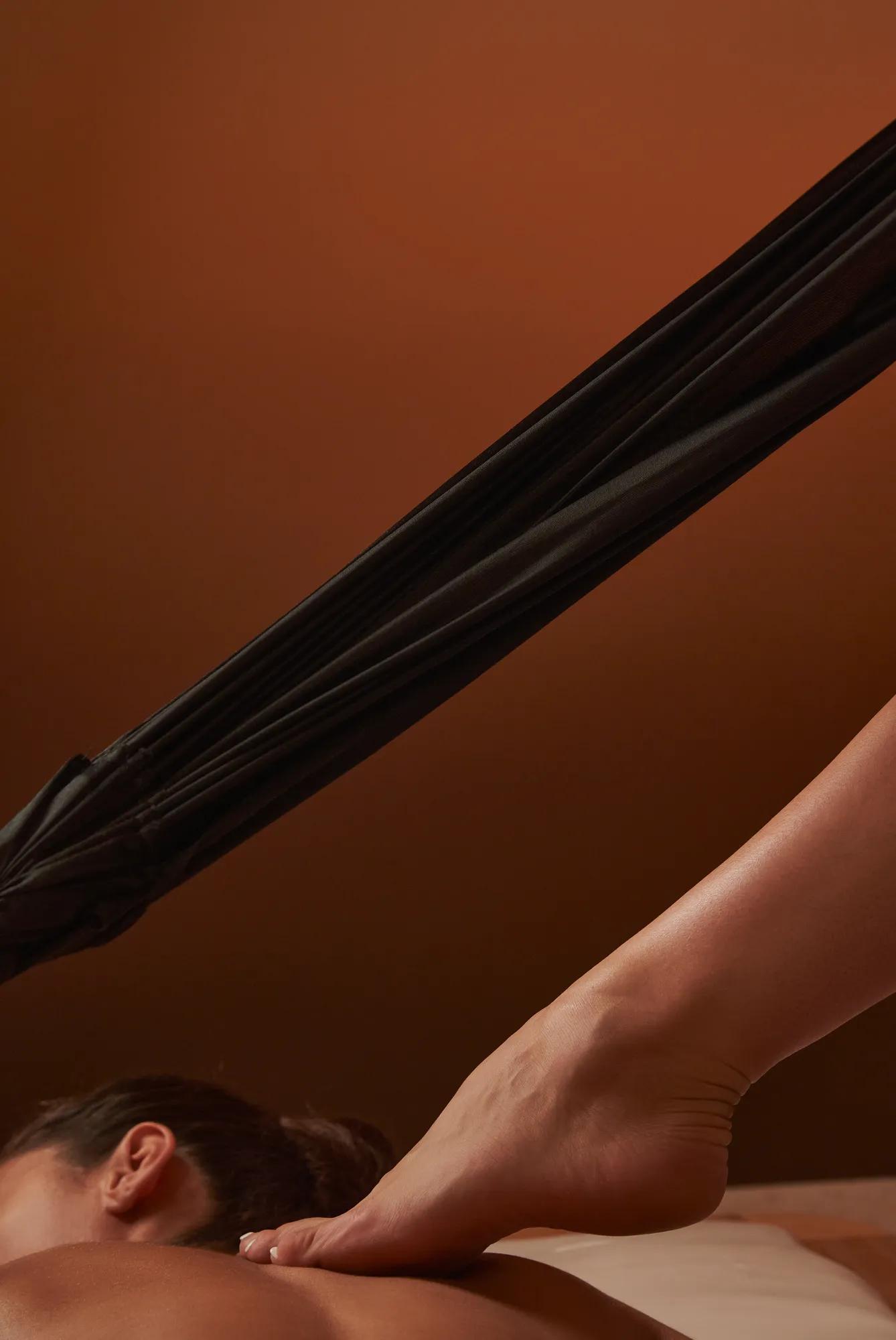 Therapist applying barefoot massage to guest’s upper back with suspended fabric support in warm spa setting.