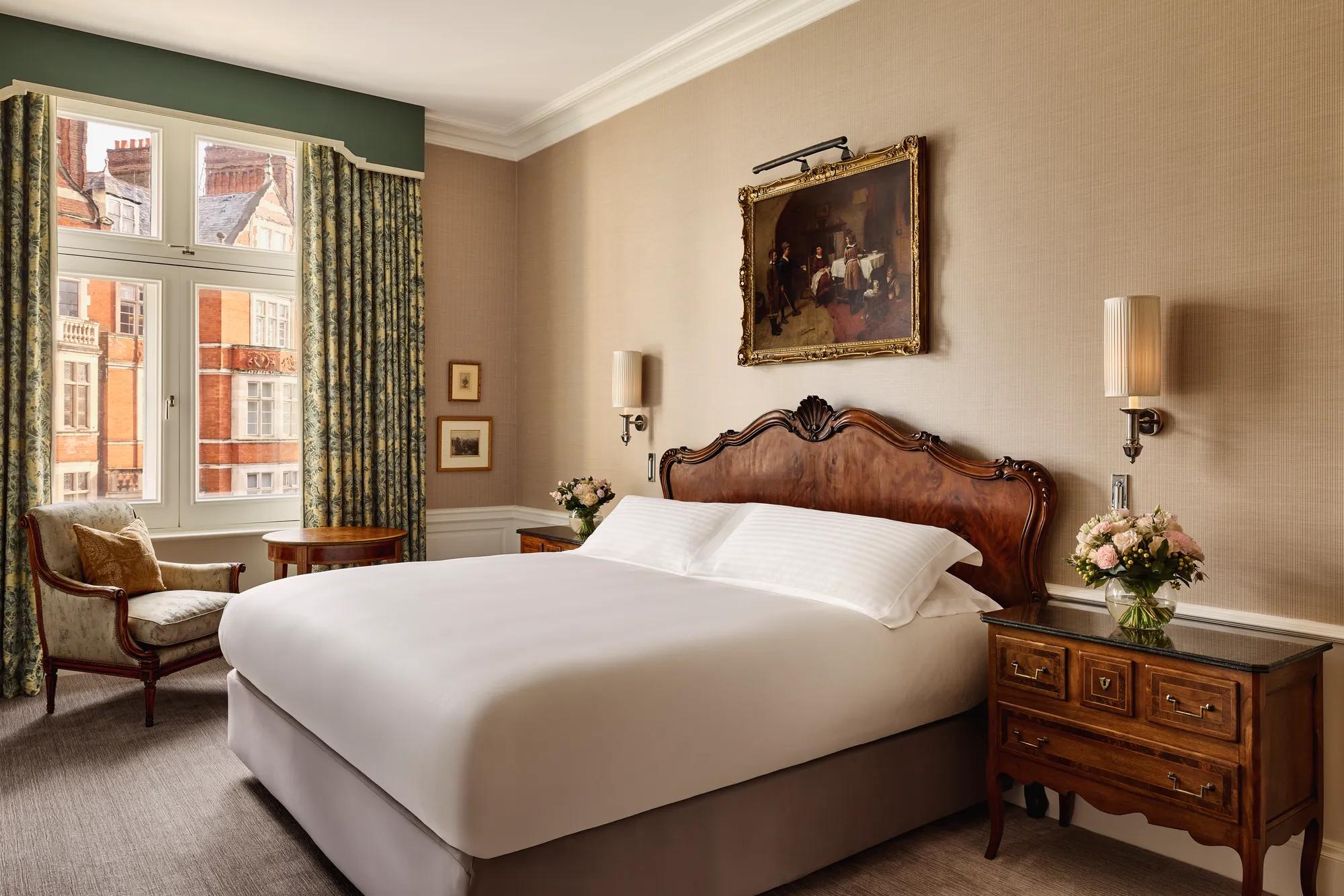 Elegant hotel bedroom with a carved wooden headboard, crisp white bedding, and antique bedside tables topped with flowers. A classical painting hangs above the bed, and natural light filters through tall windows with patterned curtains, revealing views of red-brick buildings outside.