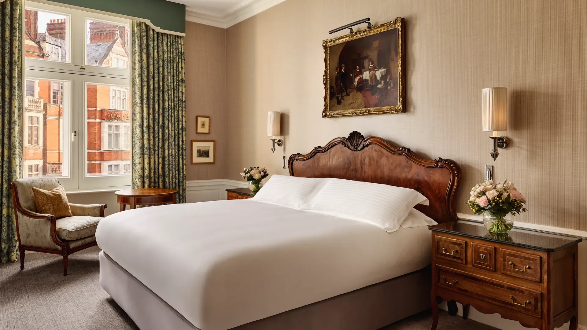 Elegant hotel bedroom with a carved wooden headboard, crisp white bedding, and antique bedside tables topped with flowers. A classical painting hangs above the bed, and natural light filters through tall windows with patterned curtains, revealing views of red-brick buildings outside.