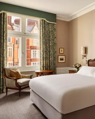 Elegant hotel bedroom with a carved wooden headboard, crisp white bedding, and antique bedside tables topped with flowers. A classical painting hangs above the bed, and natural light filters through tall windows with patterned curtains, revealing views of red-brick buildings outside.