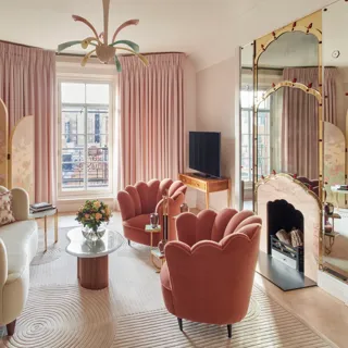 Art Deco-inspired sitting area with two coral velvet scallop armchairs and a marble coffee table, set before a decorative mirrored fireplace framed with painted detailing and flanked by colourful artwork.