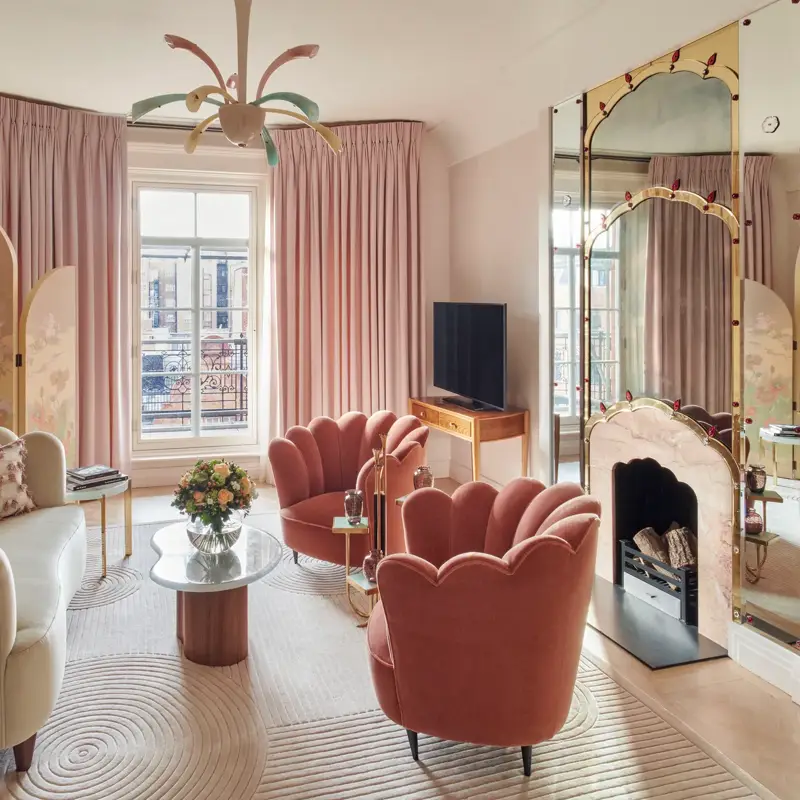 Art Deco-inspired sitting area with two coral velvet scallop armchairs and a marble coffee table, set before a decorative mirrored fireplace framed with painted detailing and flanked by colourful artwork.