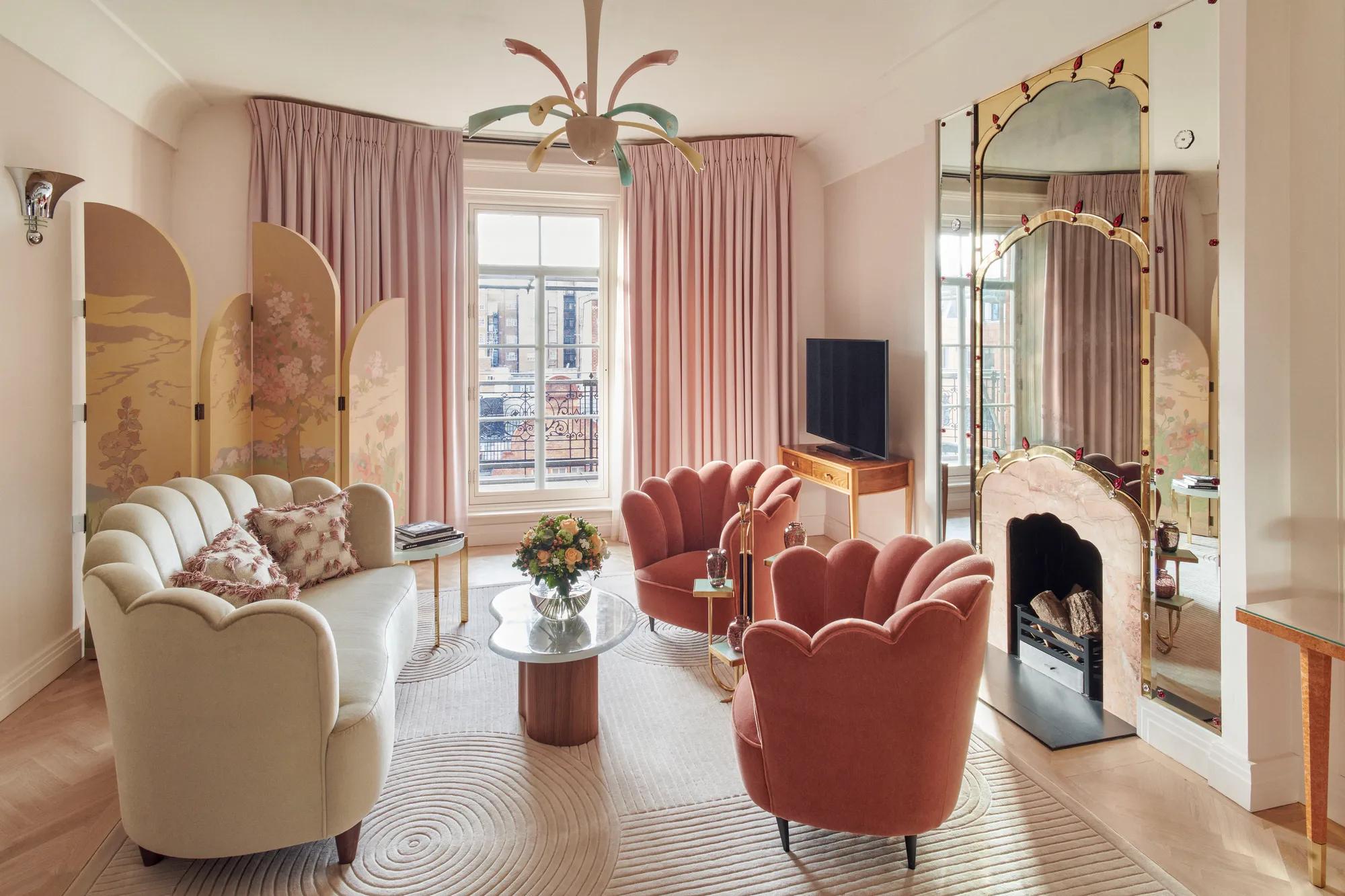 Art Deco-inspired sitting area with two coral velvet scallop armchairs and a marble coffee table, set before a decorative mirrored fireplace framed with painted detailing and flanked by colourful artwork.