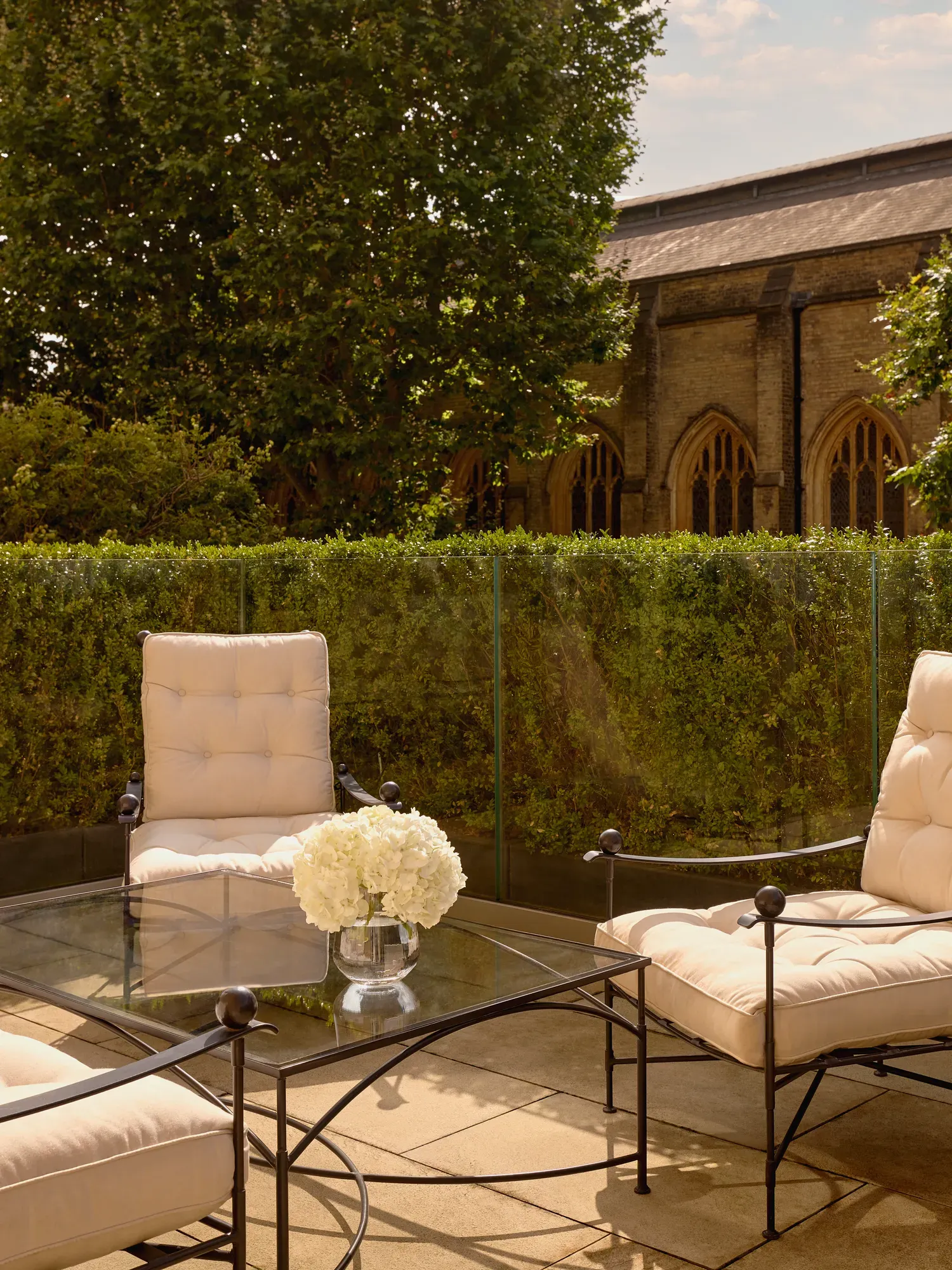 Private terrace with cushioned iron chairs and a glass table holding white hydrangeas, surrounded by greenery and views of a historic stone building with arched windows.